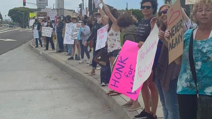 Rally attendees holding signs, standing on sidewalk along busy street