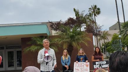 Asm. Muratsuchi holding megaphone, speaking to attendees, with students in background
