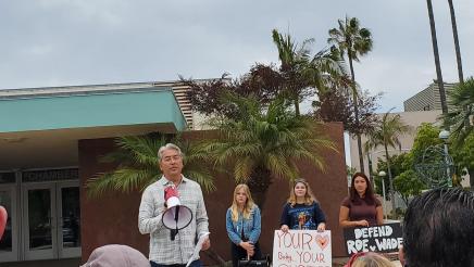 Asm. Muratsuchi holding megaphone, speaking to attendees, with students in background