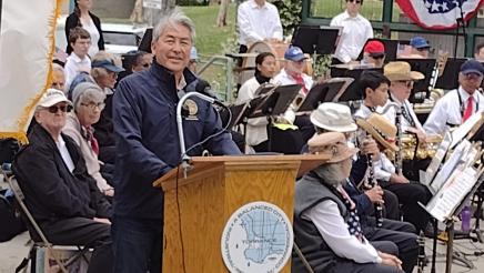 Asm. Muratsuchi on stage at podium, with band in background, speaking