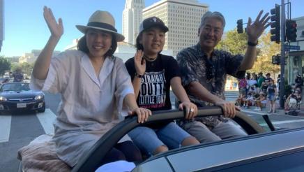 Assemblymember Muratsuchi waves at crowd from a float at a parade in downtown Los Angeles with City Hall in the background. 