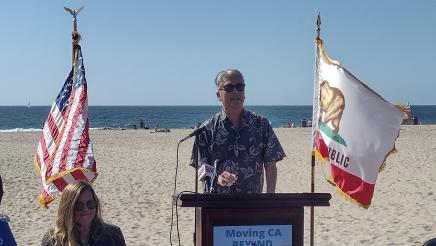 Asm. Muratsuchi at podium, speaking, with beach in background