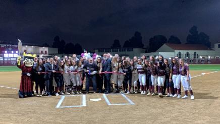 Group photo with softball players and volunteers, with ribbon being cut at center