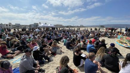 Many attendees sitting on on the beach, listening on