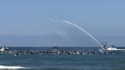 Many surfers sitting on surfboards in the ocean, escorted by lifeguard boats