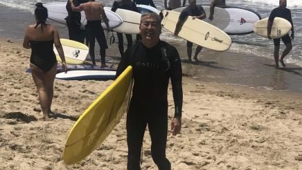 Asm. Muratsuchi in wetsuit, holding surfboard on beach, with other surfers in background