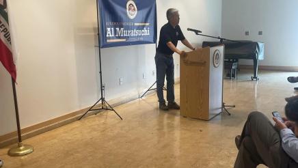 Asm. Muratsuchi at podium, speaking to audience, with large banner behind him