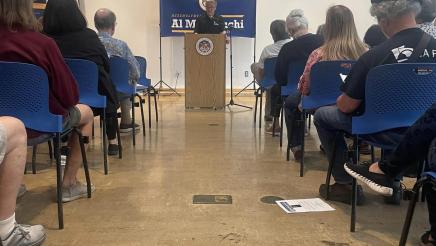 View down aisle of Asm. Muratsuchi at podium, speaking to audience, with large banner behind him