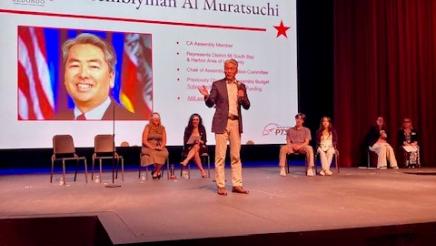 Asm. Muratsuchi onstage, speaking, with other panelists and large screen in background