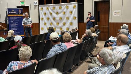 Asm. Muratsuchi standing at head of room with banner at left, speaking to attendees