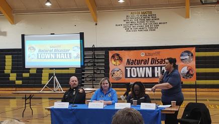 Panelists seated at table, one standing with microphone and speaking
