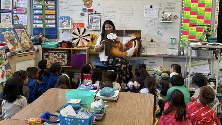 Teacher sitting in chair, reading to elementary school students, sitting on floor, in classroom