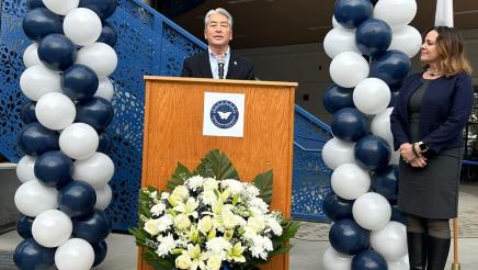 Asm. Muratsuchi at podium, in front of building, with ballon columns at left and right as a guest speaker listens on