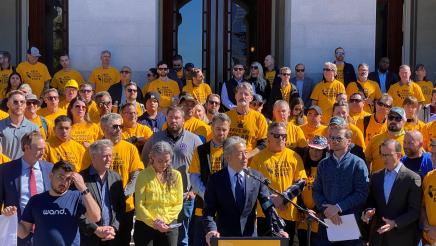 Asm. Muratsuchi at podium, speaking, with multiple participants in background wearing yellow shirts