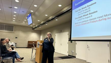 Guest speaker holding microphone, speaking to audience, with large slideshow on screen at right