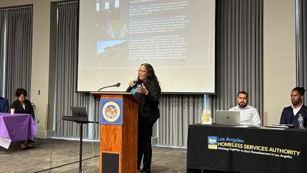 Guest speaker at podium, speaking, with slideshow on large screen in background