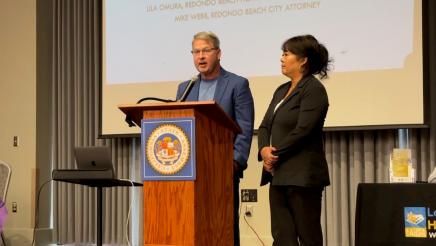 Two guest speakers at podium, one speaking, with slideshow on large screen in background