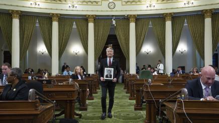 Asm. Muratsuchi walking down aisle on the Assembly Floor, holding framed photo of Alan Nishio