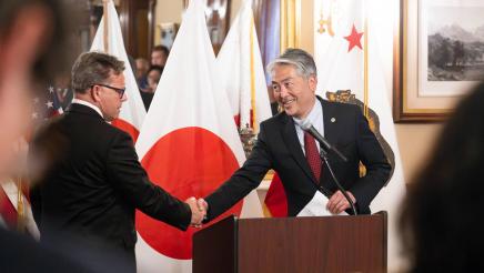 Asm. Muratsuchi at podium, shaking hands with senator, with Japanese and CA flags in background