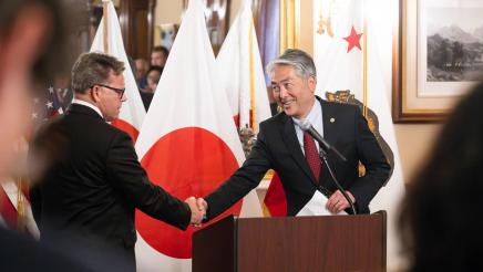Asm. Muratsuchi at podium, shaking hands with senator, with Japanese and CA flags in background