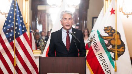 Asm. Muratsuchi at podium, speaking, flanked by U.S. and CA flags