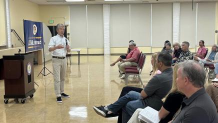 Side view of Asm. Muratsuchi standing at head of room, speaking into microphone seated attendees