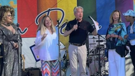 Asm. Muratsuchi standing at microphone onstage, speaking with rainbow banner in background