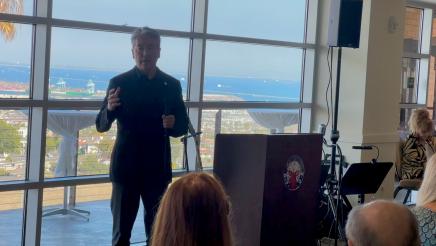 Asm. Muratsuchi standing and speaking, with ocean view through windows in background