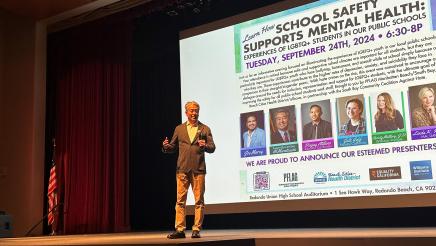 Asm. Muratsuchi standing onstage, holding microphone and speaking, with event graphic on large screen in background