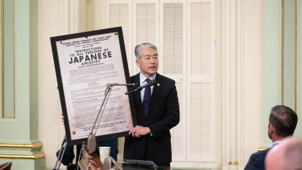 Asm. Muratsuchi holding up large framed WWII-era poster and speaking