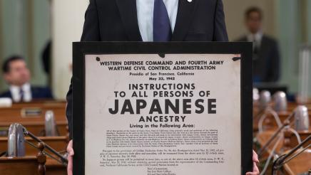 Asm. Muratsuchi holding up large framed WWII-era poster concerning Japanese citizens