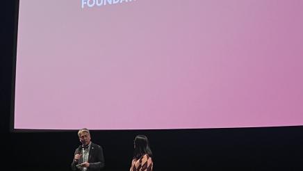 Asm. Muratsuch onstage, speaking with a fellow guest speaker, with large screen overhead