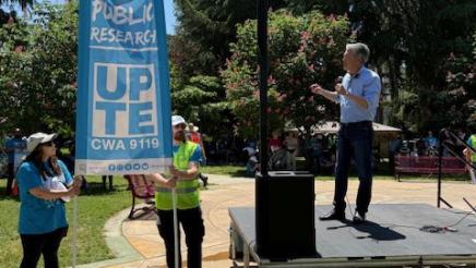 Asm. Muratsuchi onstage in park, speaking into microphone, as two workers hold banner