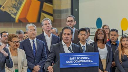 Guest speaker at podium, speaking, as Asm. Muratsuchi, Gov. Newsom and others listen on